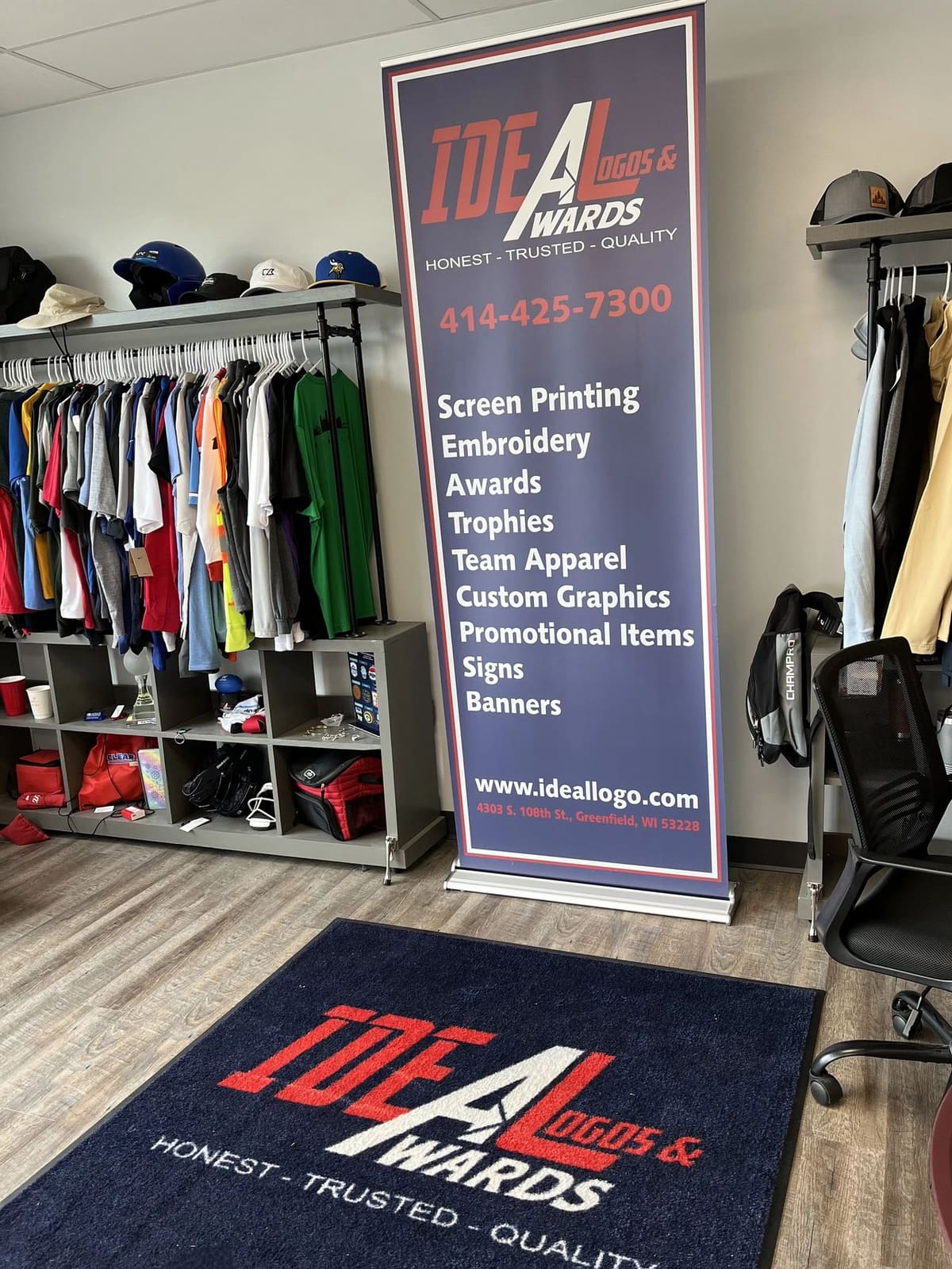 Inside the Ideal Logos and Awards shop in Greenfield, Wisconsin, with racks of custom decorated apparel, caps and a branded banner